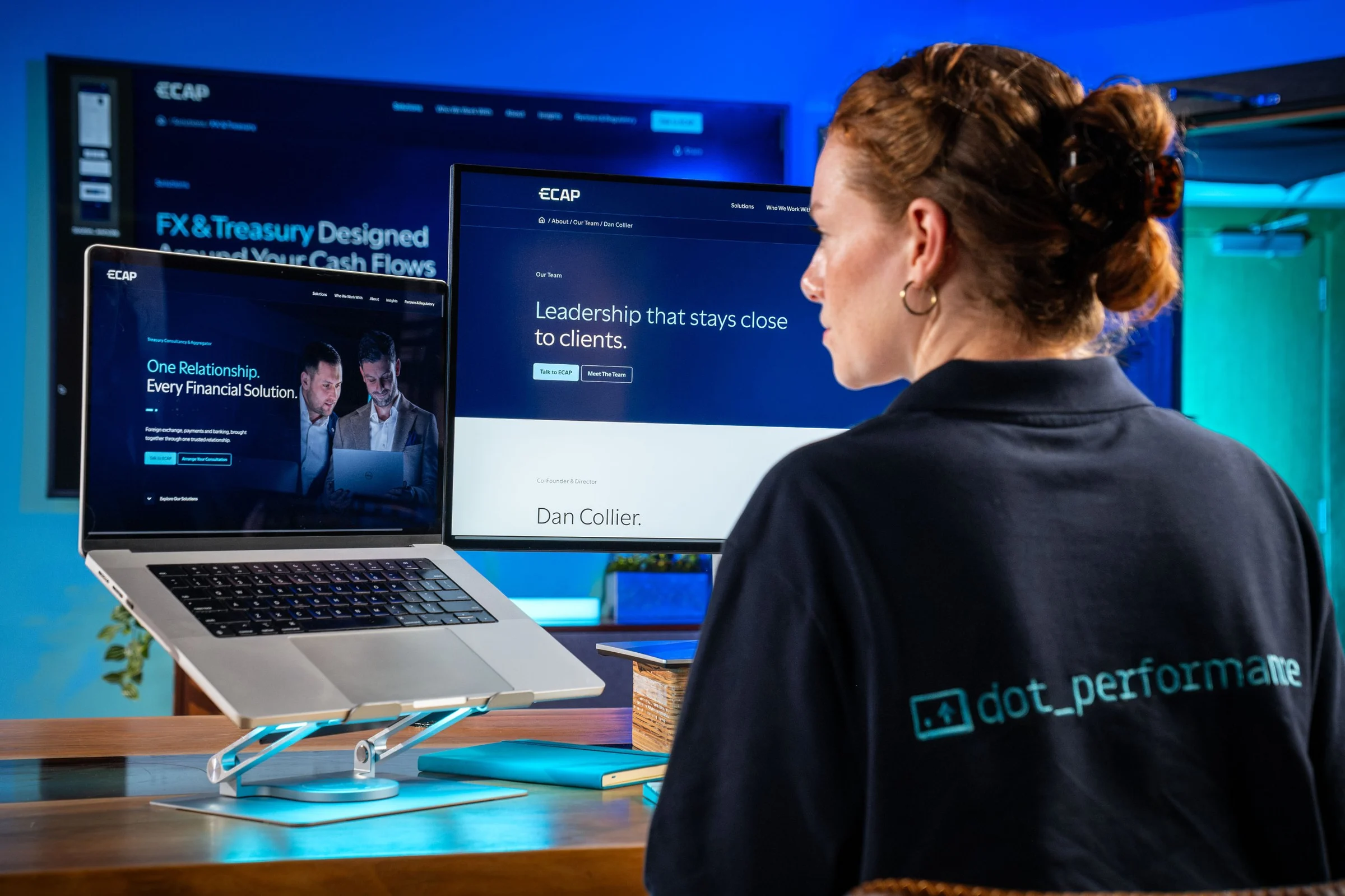 Ella Garvey, in a DotPerformance shirt, working on the ECAP website across a laptop and monitors
