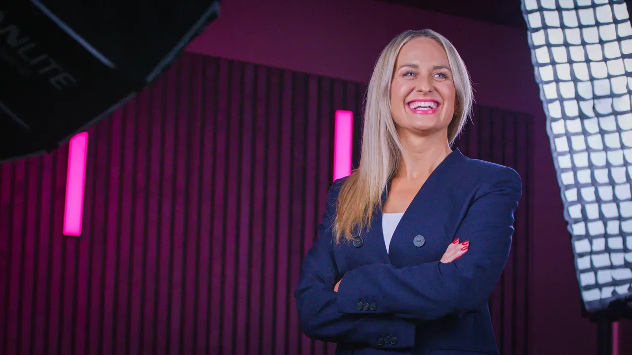 A smiling woman in a navy blazer with arms crossed against a pink striped background.