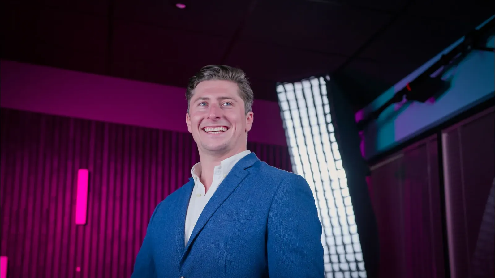 Smiling man in a blue blazer in a studio with pink and purple lighting.
