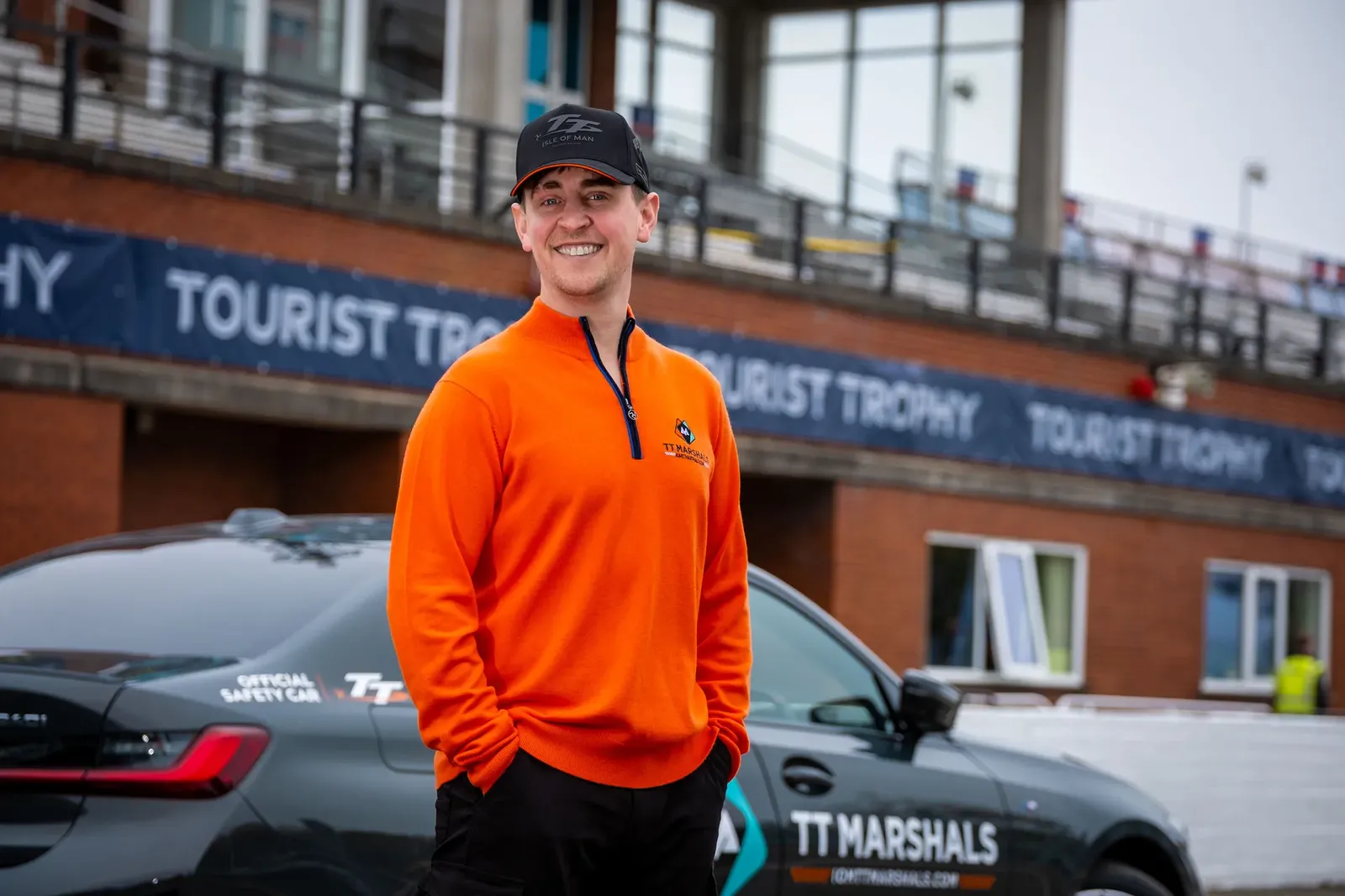 Dom Herbertson smiling in front of the Tourist Trophy Grandstand and the TT Marshals car, wearing the prize jumper