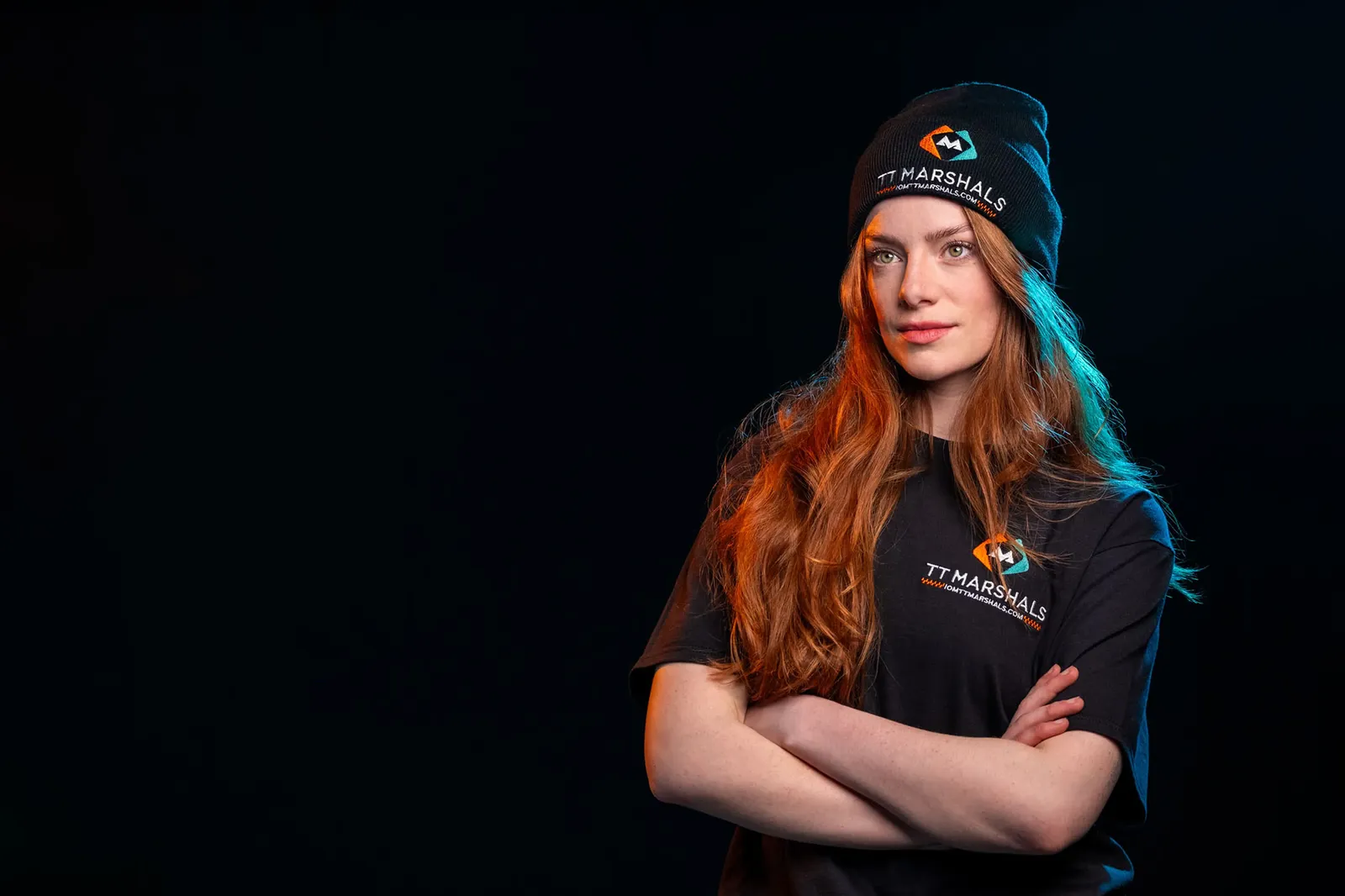 Ella Garvey in TT Marshals merchandise, arms crossed, wearing a marshals beanie and t-shirt against a black background